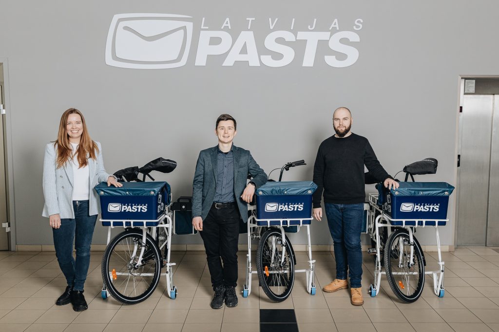 2 Mapon and 1 Latvijas Pasts employees standing next to postal e-bikes.