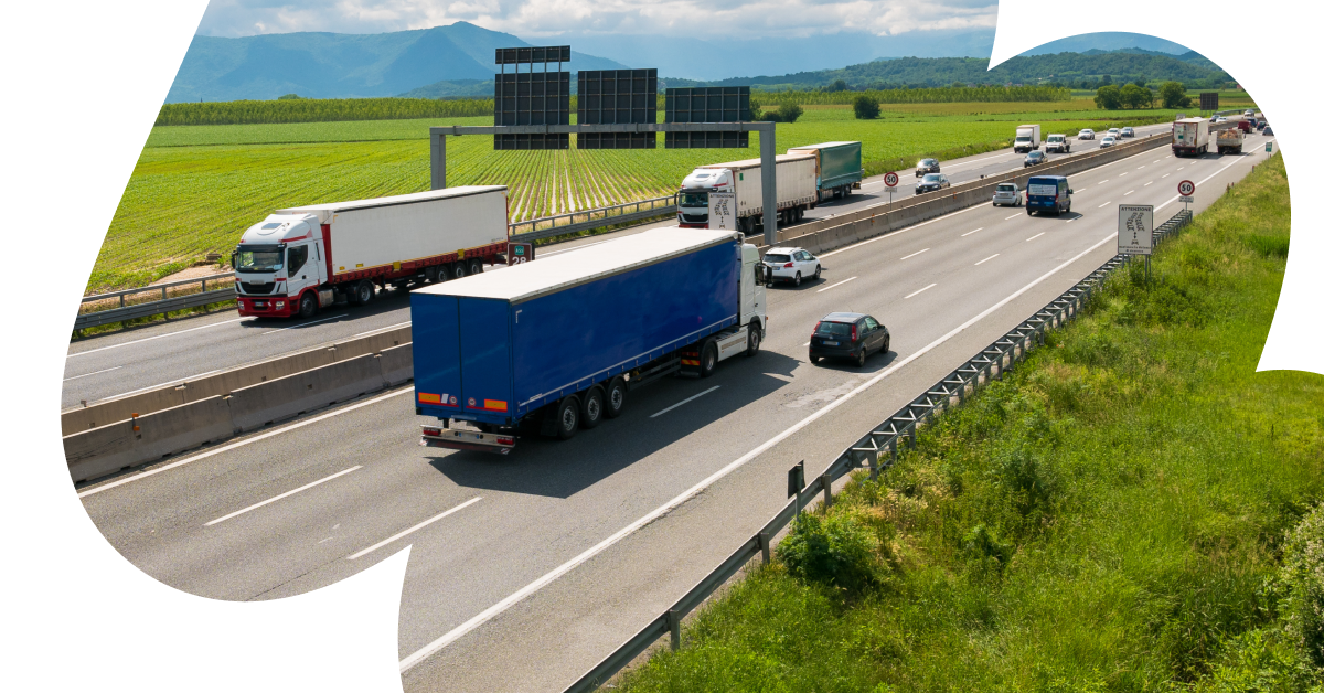 Šoseja ar vairākām joslām, kur abos virzienos pārvietojas kravas auto un vieglie automobiļi, apkārt zaļi lauki un tālumā kalni.