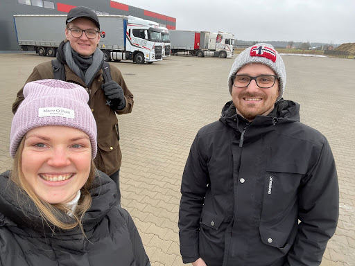 Medarbejdere fra Mapon smiler under et besøg hos RMC-Transports, med lastbiler og logistikfaciliteter i baggrunden.